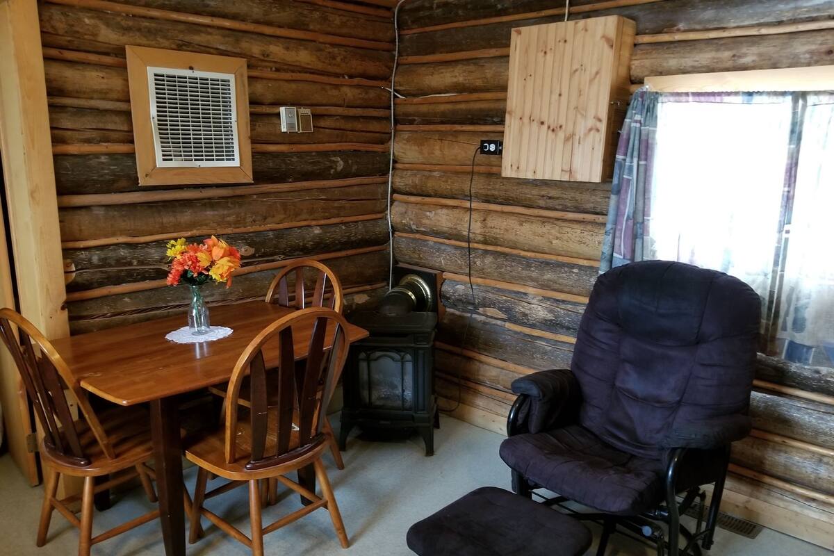 A wooden cabin interior with a dining table, chairs, a sofa, and a small heater.