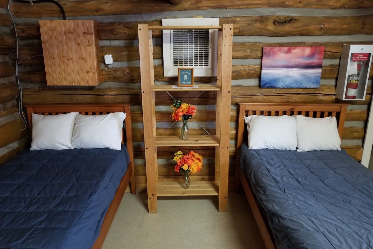 A cabin-style room with two beds, a wooden shelf with a vase of flowers, an air conditioning unit, and a painting on the wall.