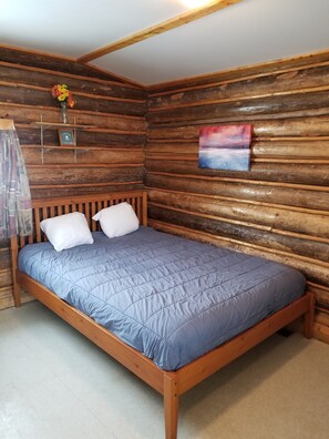 Une pièce rustique aménagée dans un chalet en bois, avec un lit, des oreillers, un vase rempli de fleurs, un tableau encadré et une étagère sur laquelle se trouvent une bouteille en verre et une petite boîte.