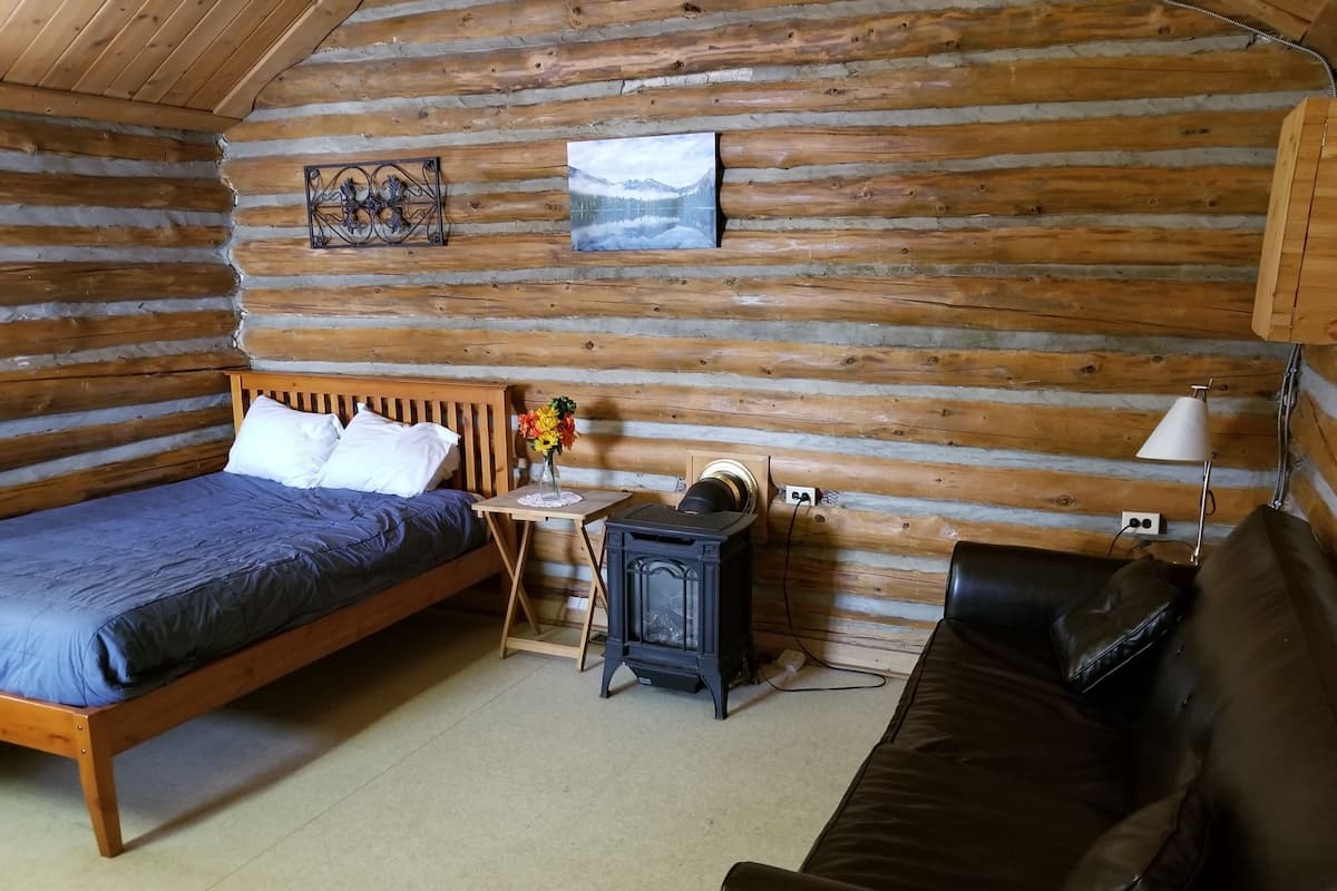 A log cabin room with a wooden bed, a small table with a vase of flowers, a black stove, and a leather couch.