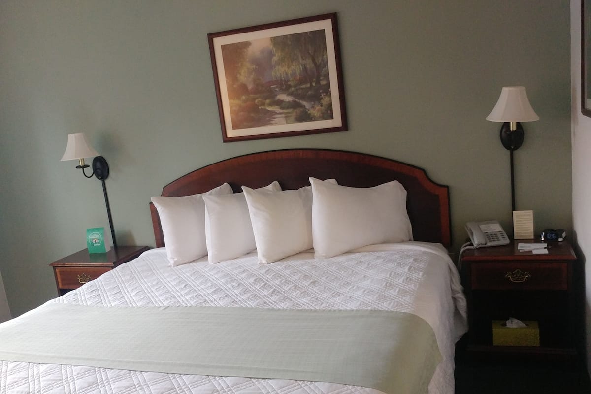 A neatly made bed with a quilted white comforter and pillows, a framed painting above the headboard, and two bedside tables with lamps and a phone.
