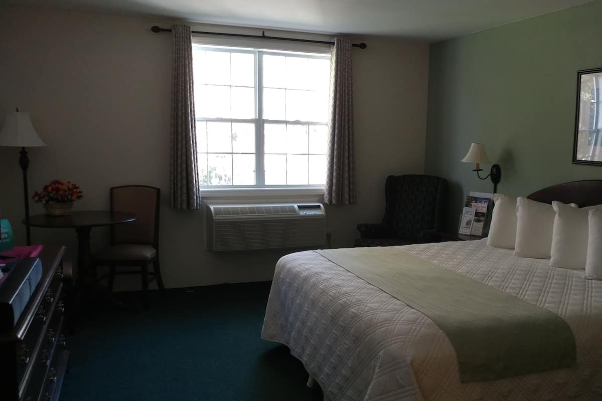 A hotel room with a large bed, a desk with a chair, a window with curtains, an air conditioning unit, and a framed picture on the wall.