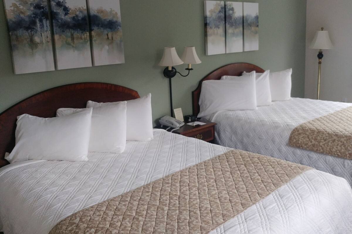 Two double beds with white linens and a quilted beige throw blanket, a bedside table with a lamp, and two wall-mounted paintings.