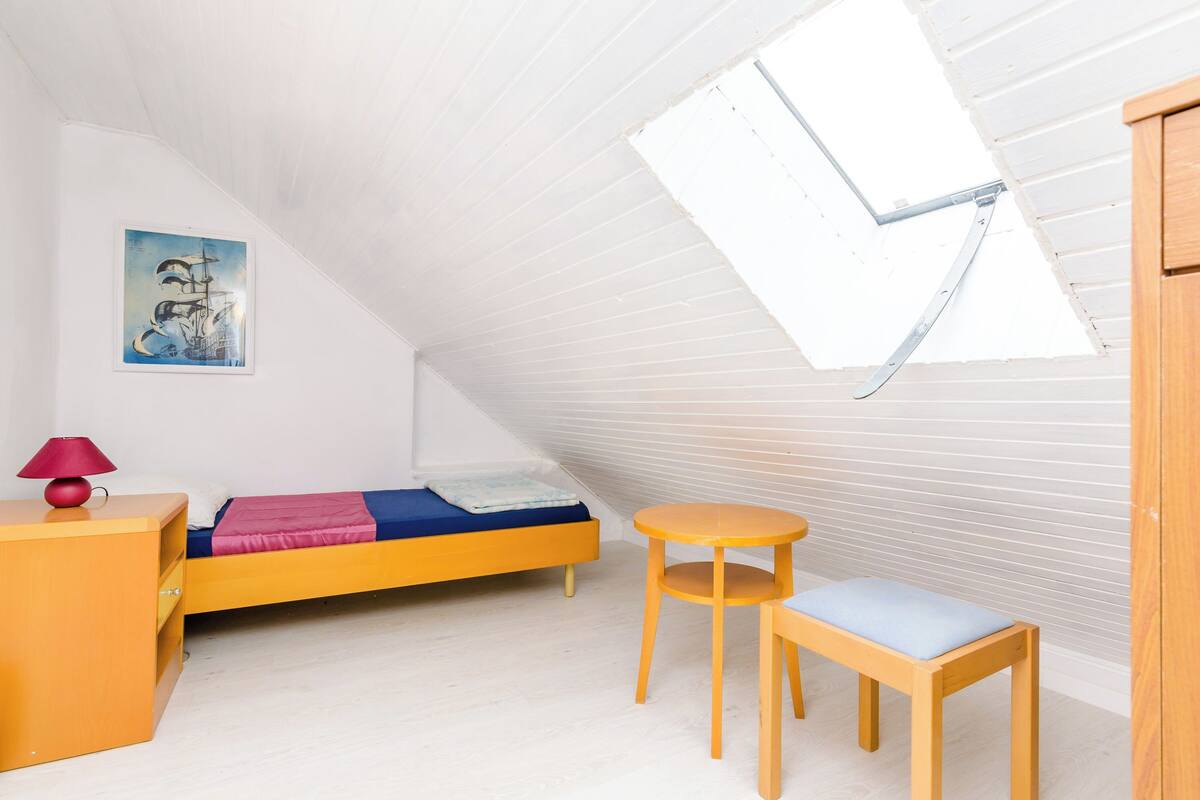 Un pequeño y luminoso dormitorio en el ático, con techo inclinado, una cama individual, una mesita, una silla y una lámpara roja.