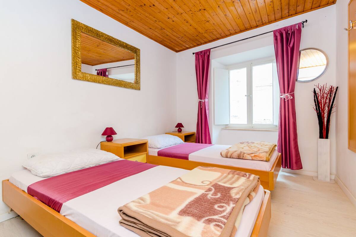 Una habitación con dos camas, cabeceros de madera, un espejo, una ventana con cortinas, una mesita de noche con una lámpara y un jarrón con ramas decorativas.