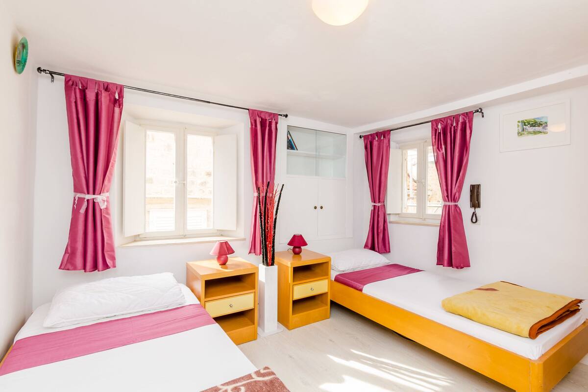 Una habitación con dos camas, una mesita de noche de madera, un espejo y cortinas rosadas.