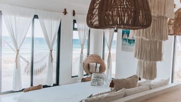 Una habitación frente a la playa con cama, ventanas amplias y una silla de mimbre.