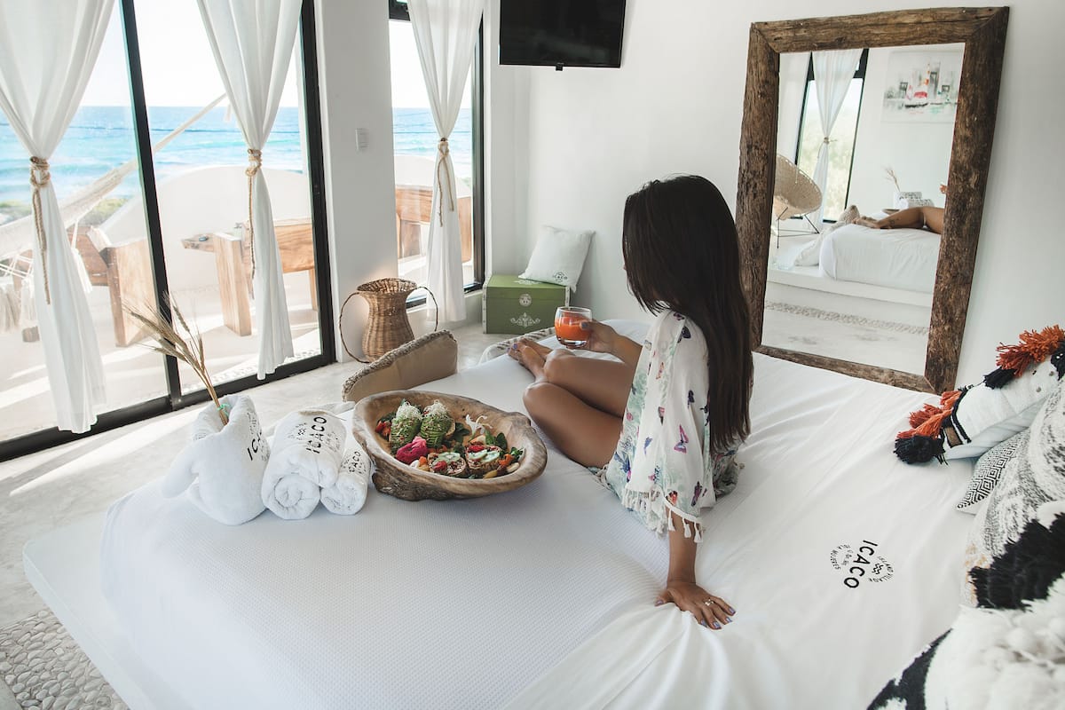 A bedroom with a large window, a bed, a bowl of fruit, and a mirror.