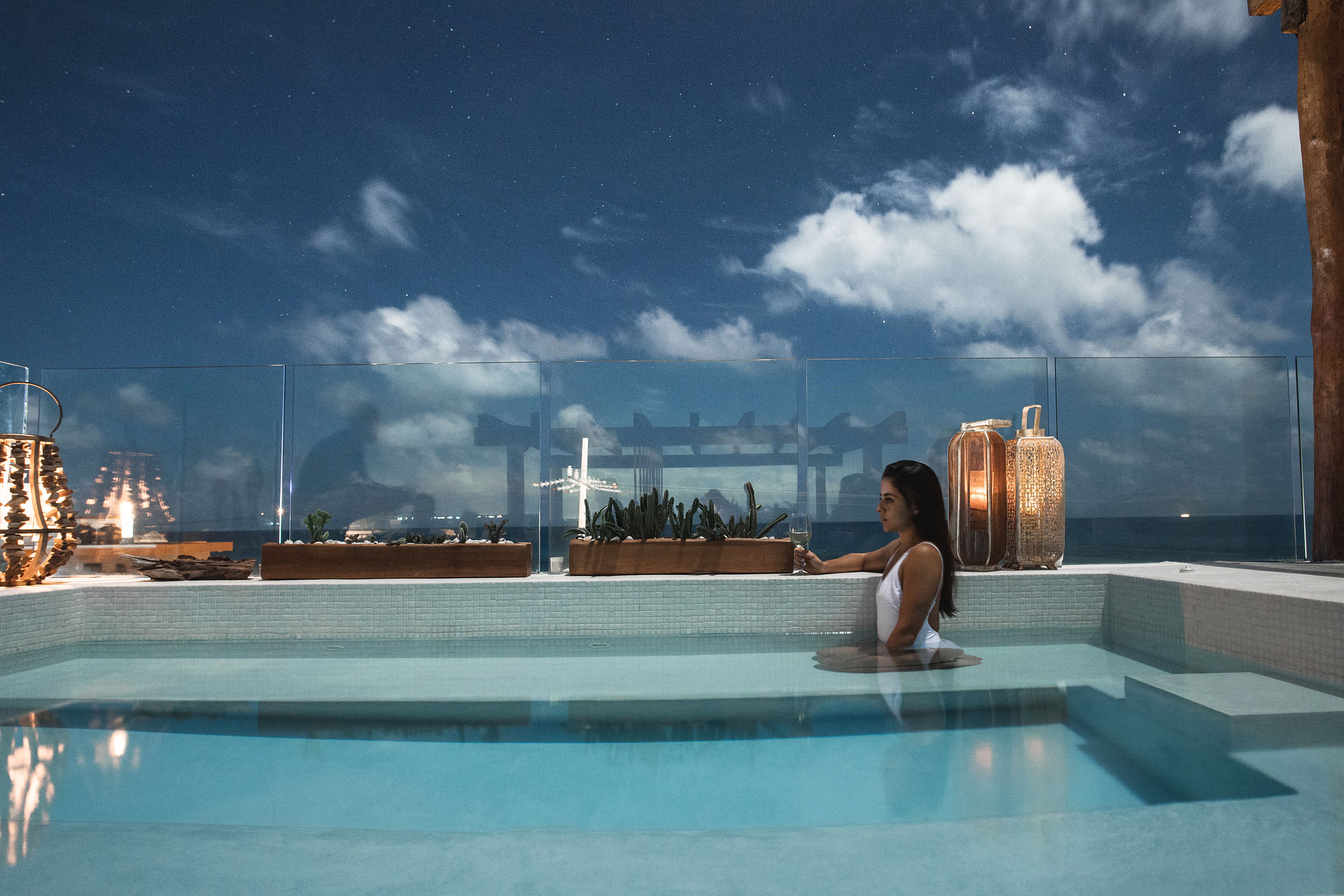 A pool area with a clear view of the night sky, a glass railing, and a person relaxing by the pool.