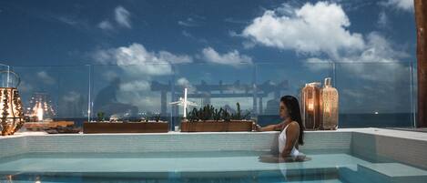 A pool area with a clear view of the night sky, a glass railing, and a person relaxing by the pool.
