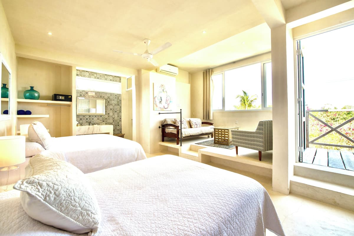 A hotel room with two beds, a sitting area with a sofa and armchairs, a ceiling fan, and a large window with a view of greenery.