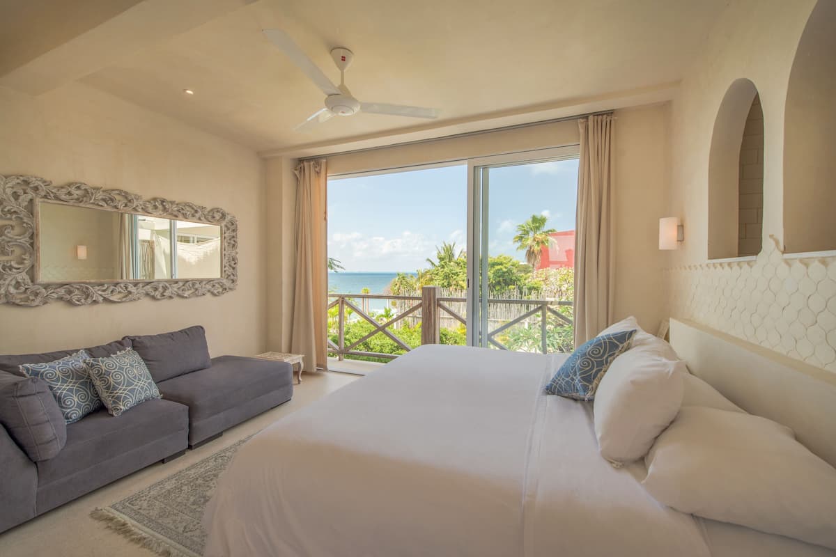 A bedroom with a large bed, a grey sofa, a ceiling fan, and a balcony with a view of the ocean.