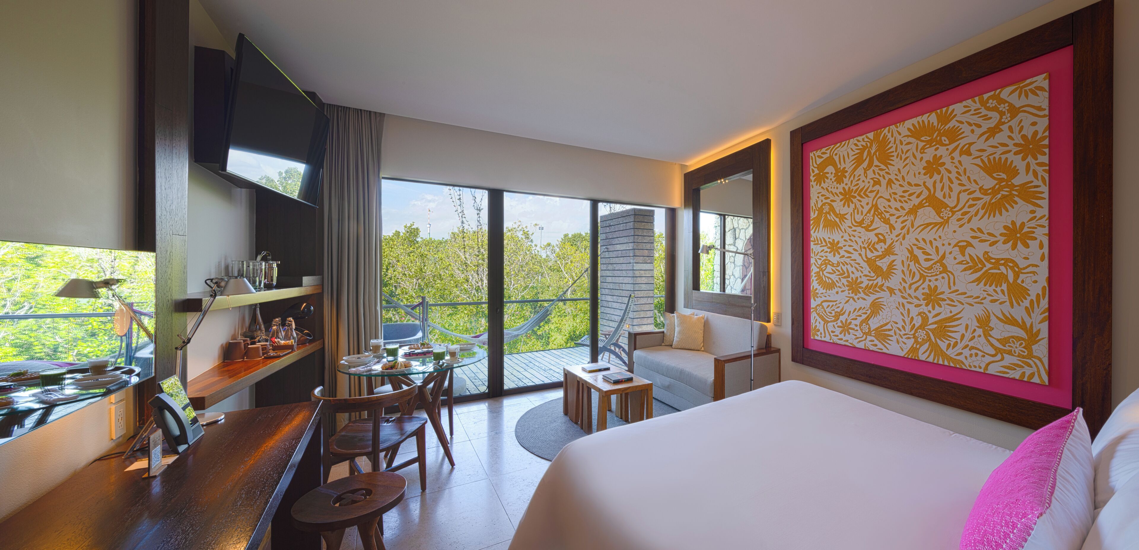 Habitación de hotel moderna con una cama grande, una zona de comedor con mesa y sillas, un televisor montado en la pared y un balcón con vistas a árboles.