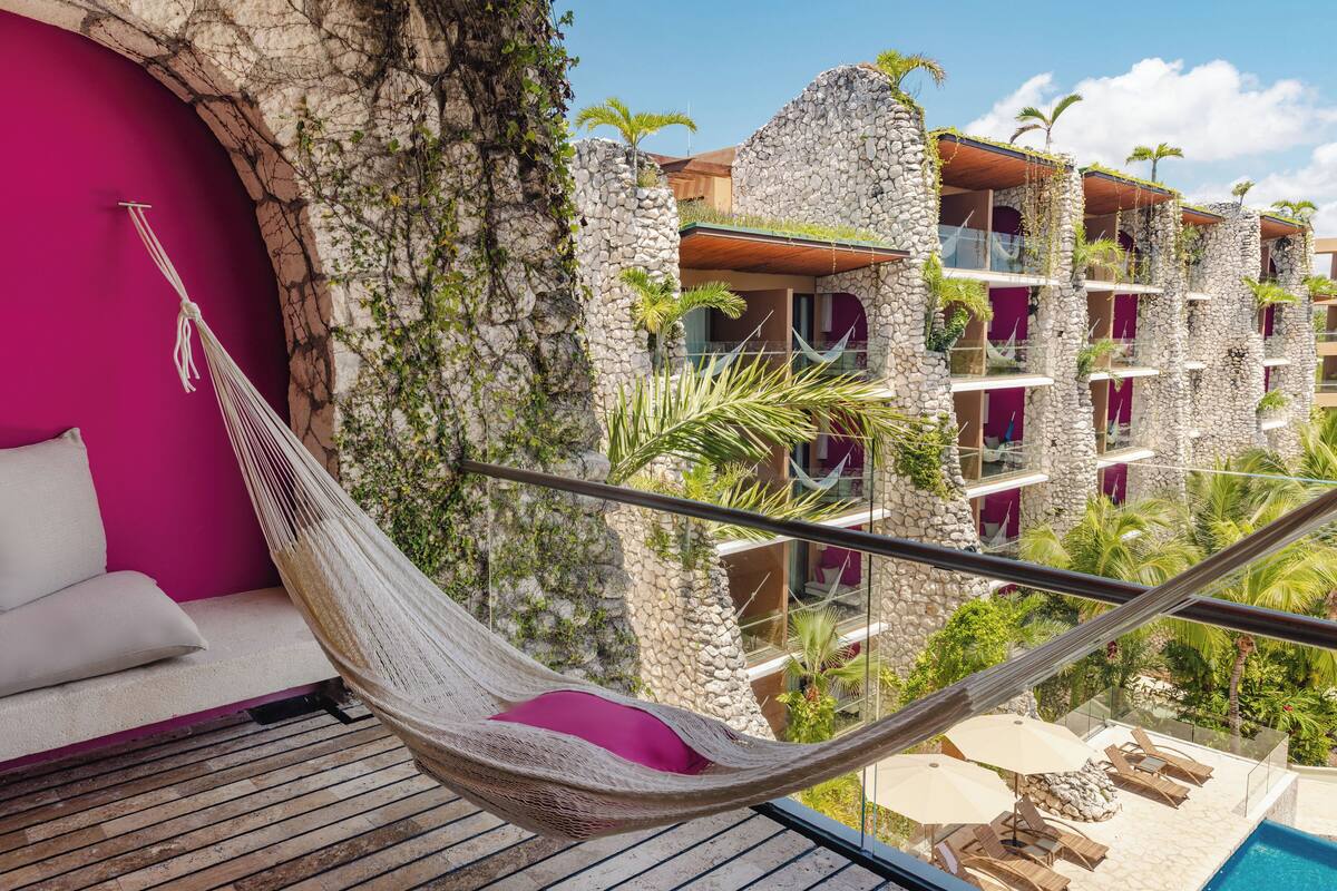 A hammock on a balcony with a view of a pool and a building with stone walls and balconies.