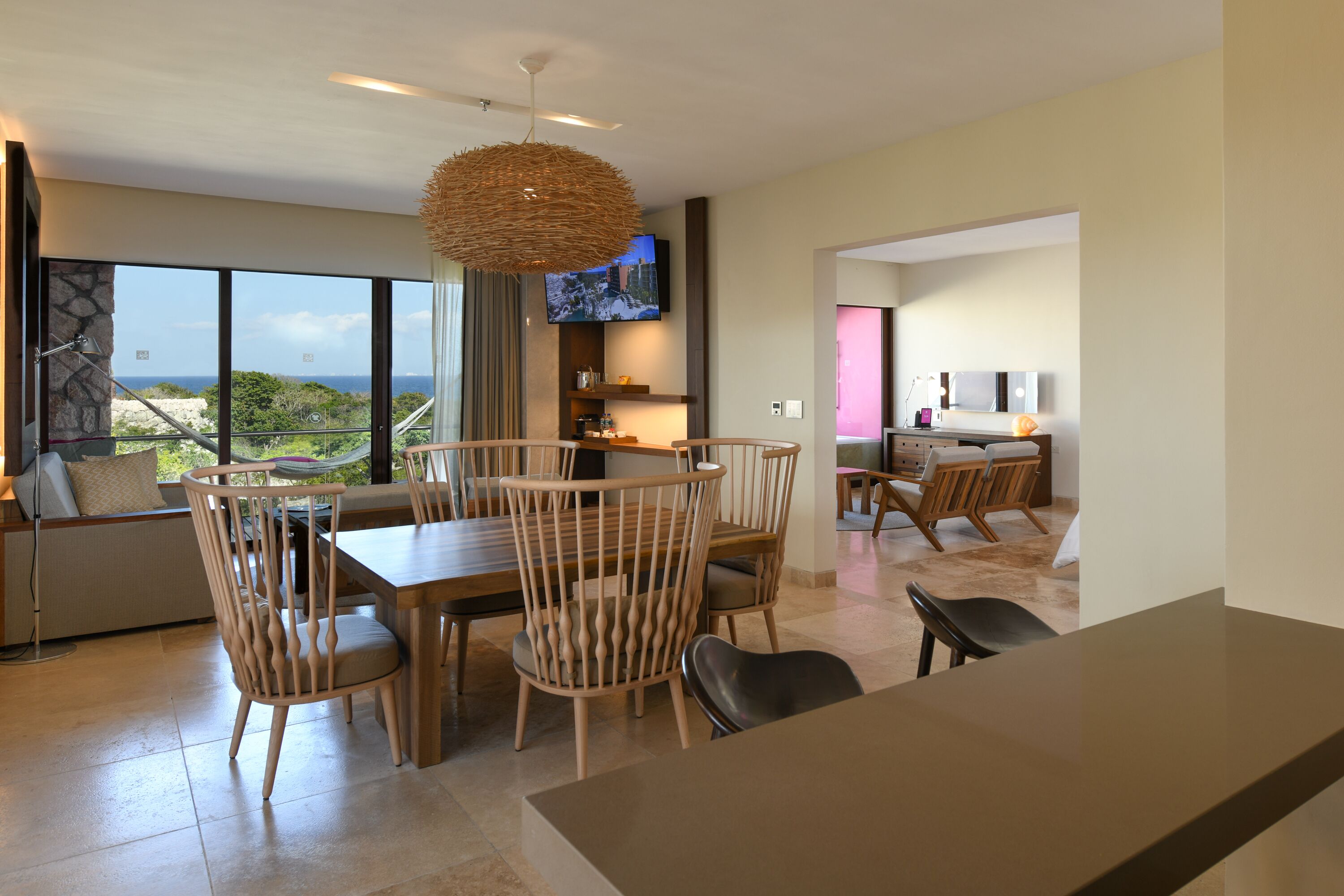 A modern dining area with a large table and chairs, a television mounted on the wall, and a view of the ocean through large windows.