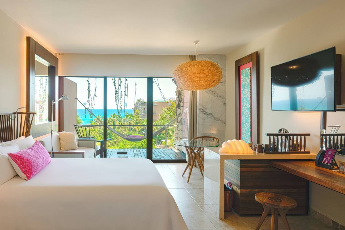 A hotel room with a bed, desk, and a TV. The room has a balcony with a view of the sea.