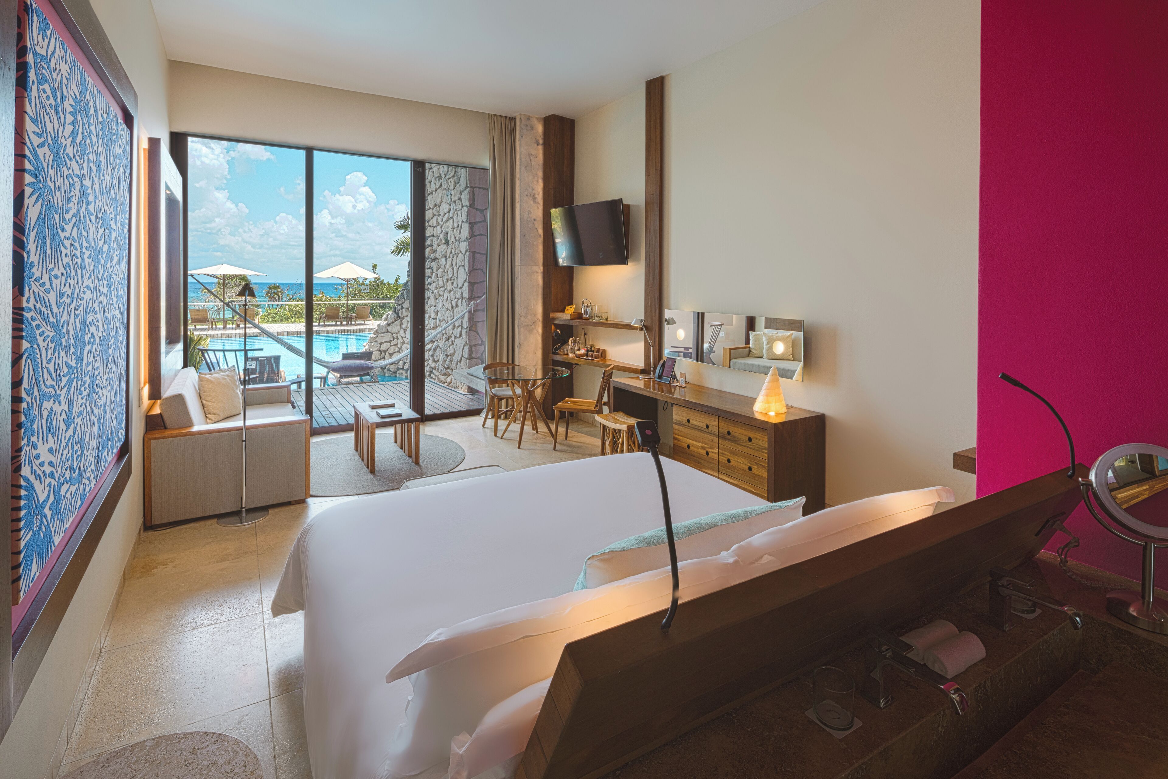 A hotel room with a large bed, a desk, a chair, a TV, and a view of a pool and palm trees.