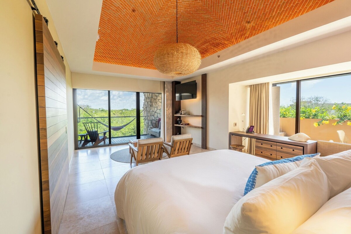 A modern hotel room with a large bed, a desk, a chair, and a balcony with a view.