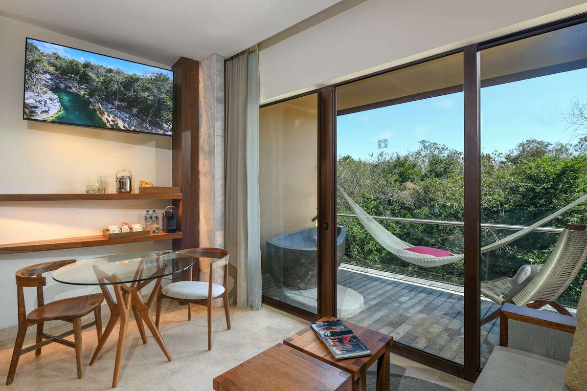A modern room with a glass table, wooden chairs, a large TV, and a balcony with a hammock.