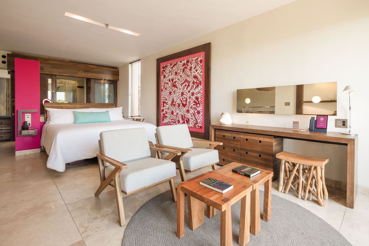 A modern hotel room with a bed, two chairs, a desk, and a small stool.