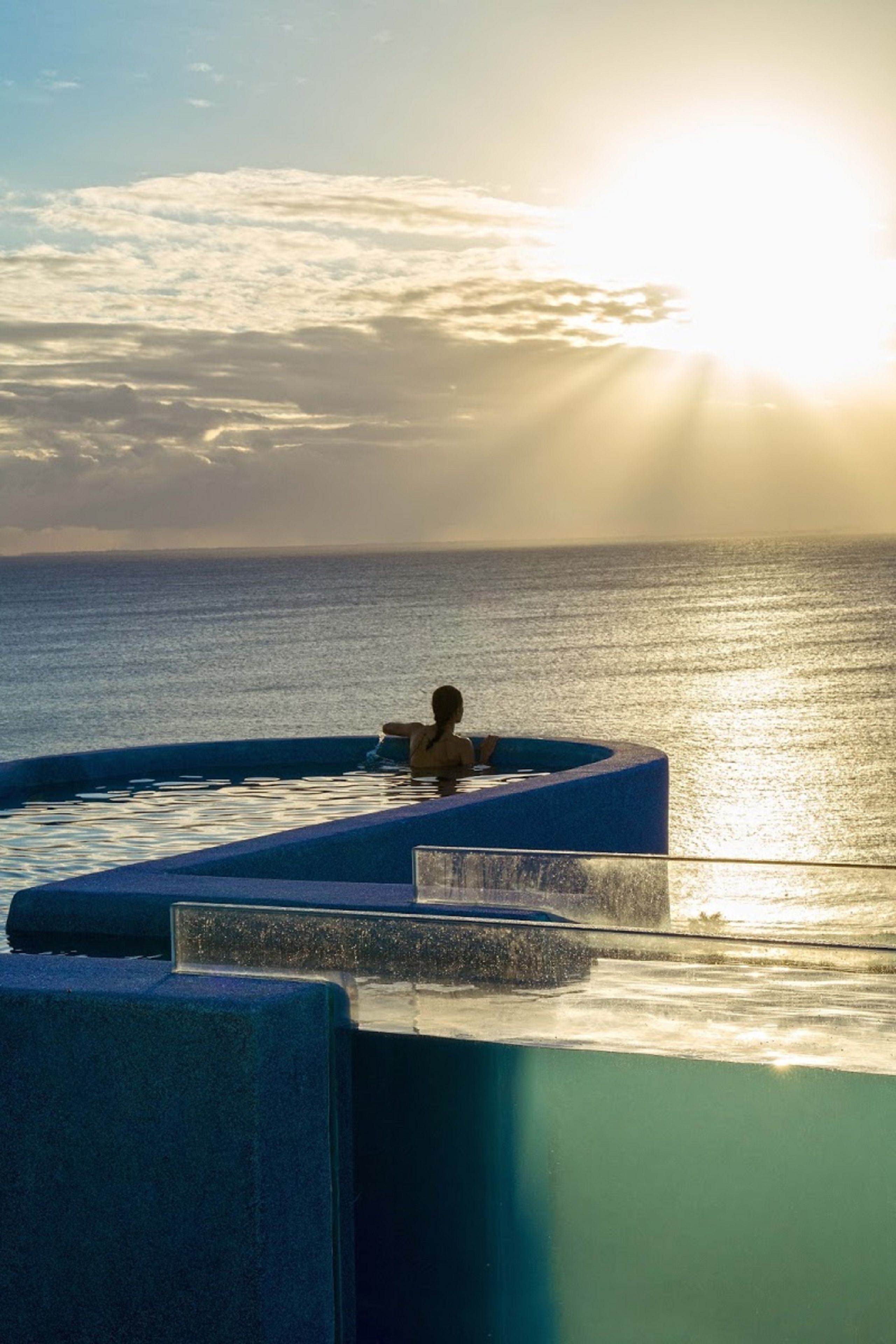 A person relaxing in a blue infinity pool with a view of the ocean and the sun setting in the background.