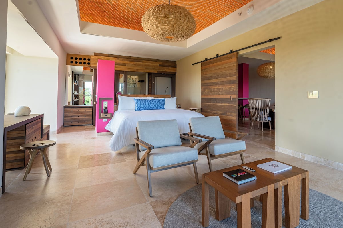 A modern bedroom with a large bed, two armchairs, a wooden coffee table, and a dining area with a chair and table.