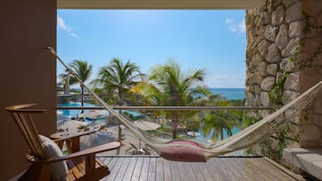 Un balcón con una hamaca, sillas de madera y vistas a una piscina y palmeras.