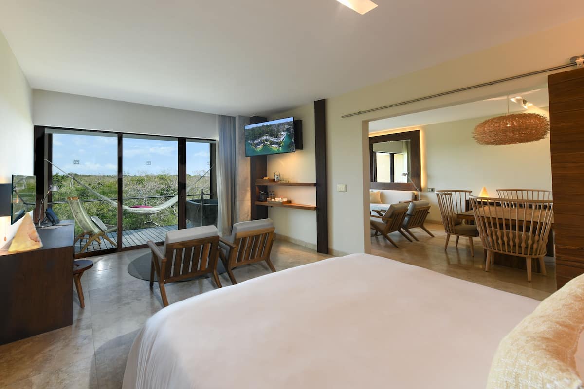 A hotel room with a large bed, a desk, a TV, a balcony with chairs, and a dining area.