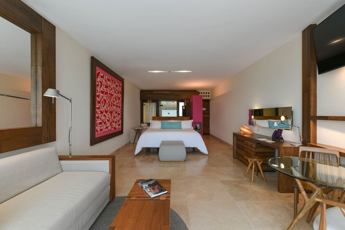 A modern hotel room with a bed, a desk, a sofa, a glass table, and a large mirror.