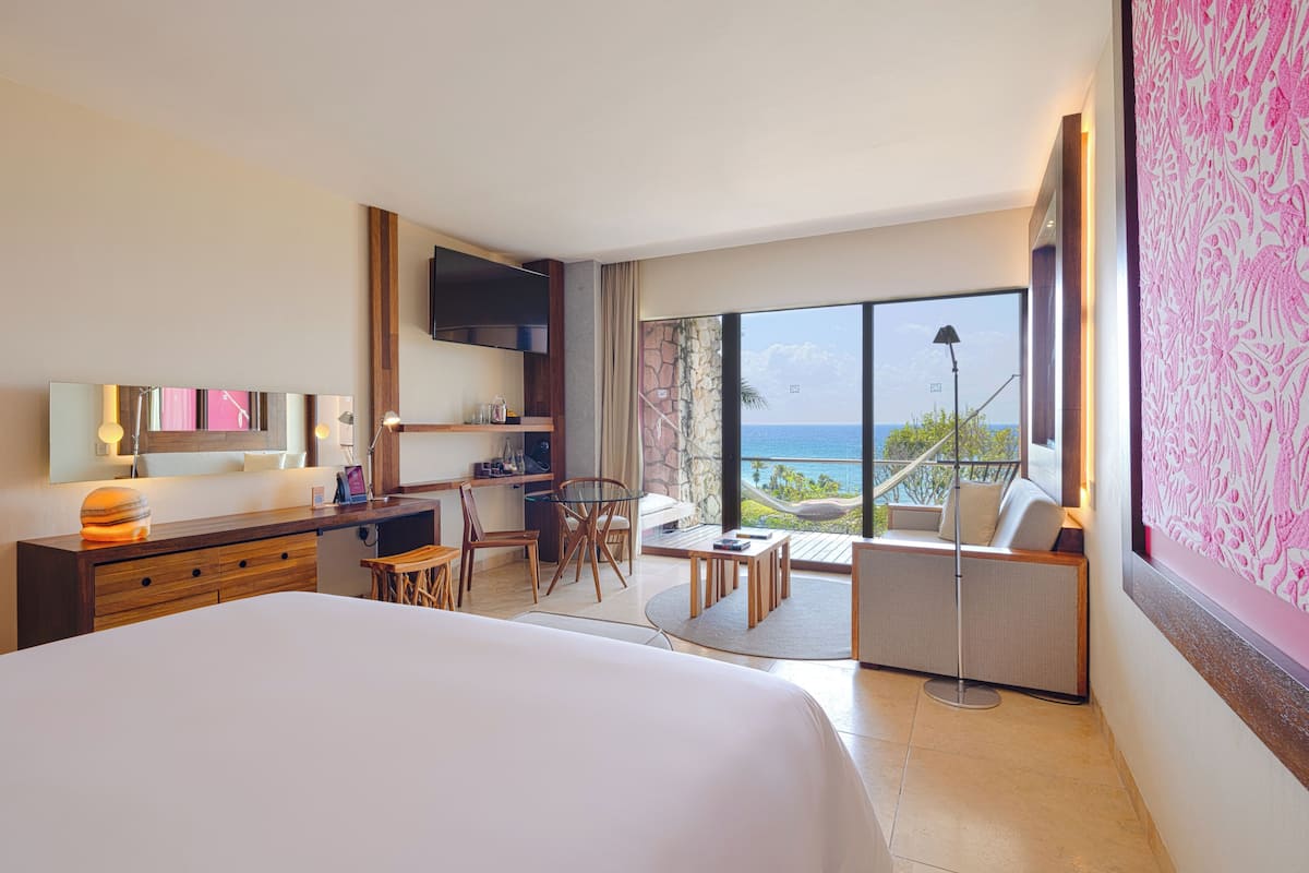 A hotel room with a large bed, a desk, a chair, a small table, and a sofa. There is a TV mounted on the wall and a window with a view of the ocean.