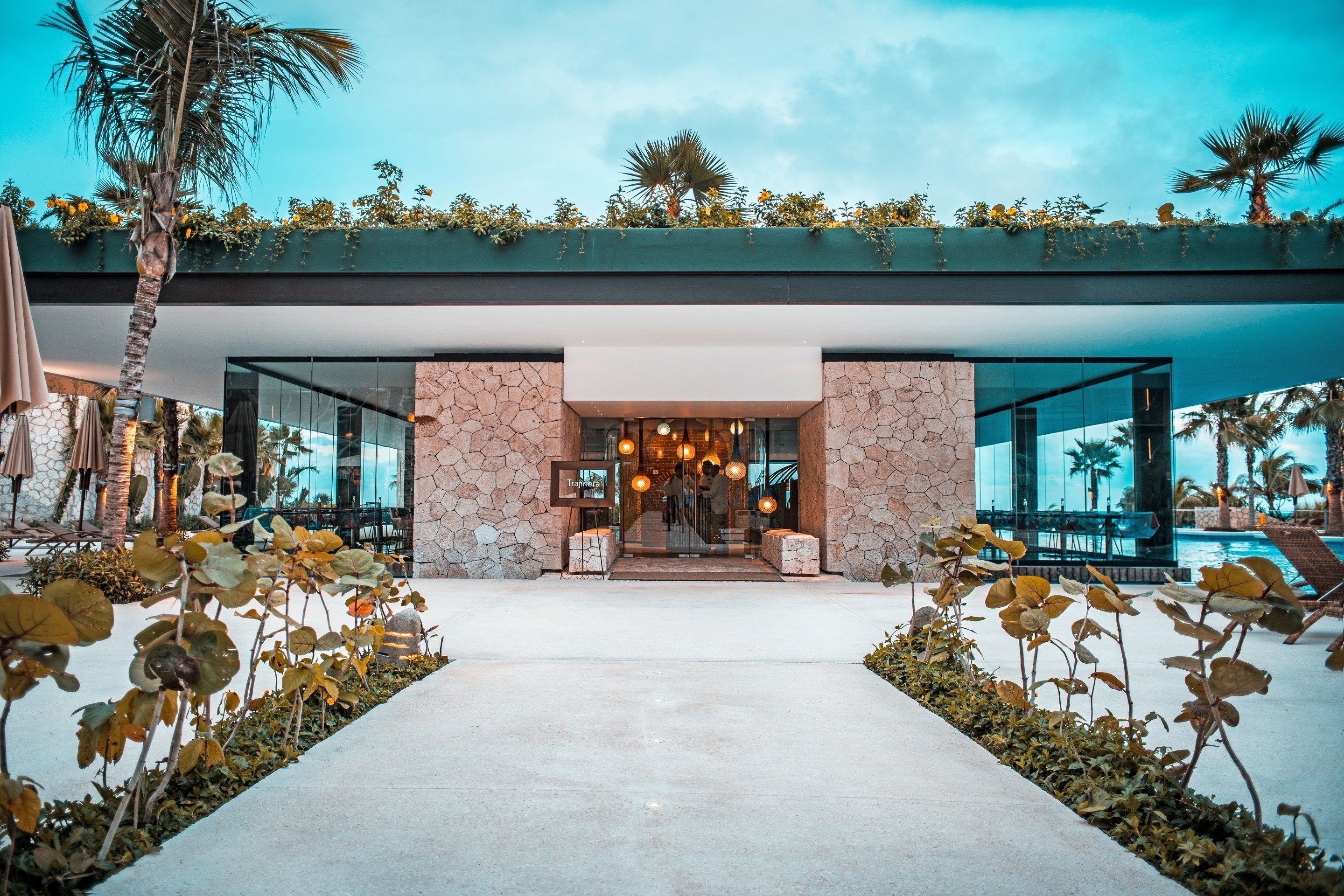 Una entrada de hotel moderna con fachada de piedra, puertas de vidrio y una piscina al fondo.