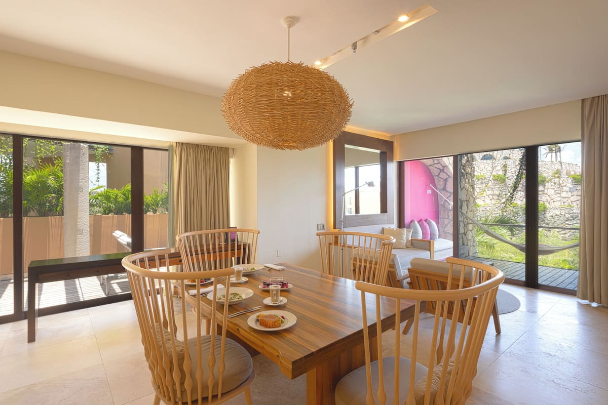 A dining area with a wooden table and chairs, a woven pendant light, and large windows offering a view of greenery.