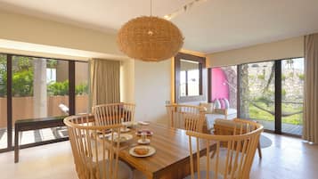 A dining area with a wooden table and chairs, a woven pendant light, and large windows offering a view of greenery.
