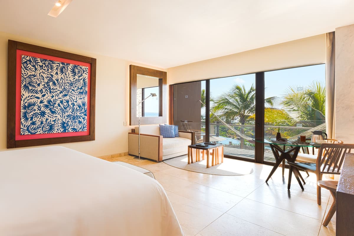 A hotel room with a large bed, a desk with a chair, a balcony with a view of palm trees, and a large framed artwork on the wall.