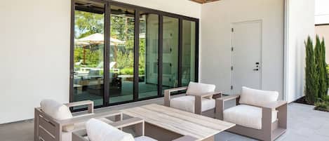 Un patio exterior con mobiliario moderno, un ventilador de techo y un gran ventanal corredizo que da a un jardín.