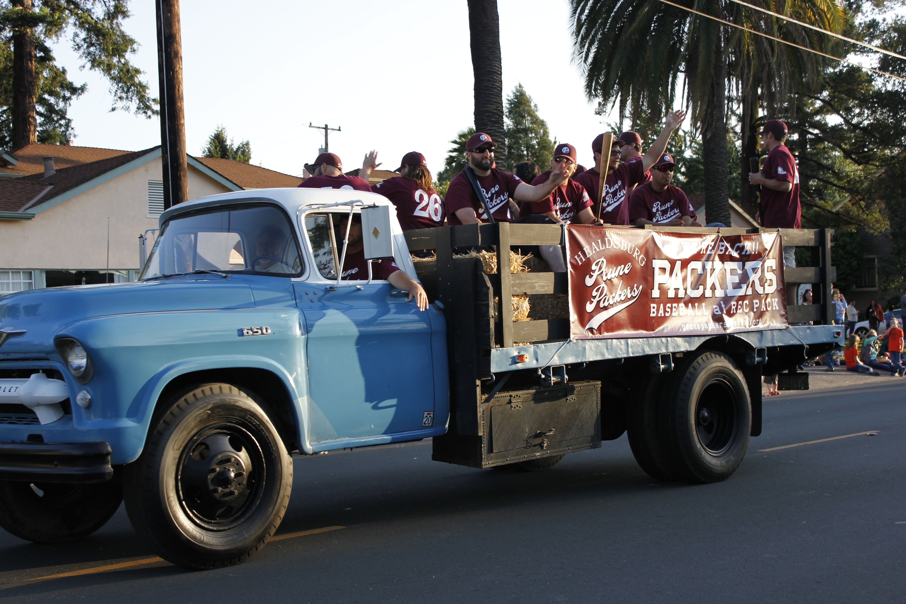 A vintage blue truck with a wooden bed carrying a group of people in maroon uniforms, possibly for a parade or event.