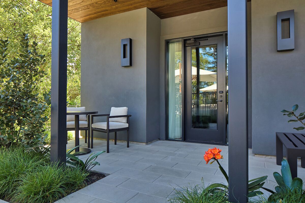 A modern patio with a table and chairs, a glass door entrance, and a small garden area with plants.