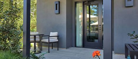 A modern patio with a table and chairs, a glass door entrance, and a small garden area with plants.