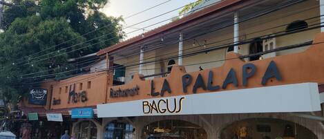 Vista de la calle con un edificio que muestra los rótulos "La Palapa" y restaurante "Bacus". Hay coches estacionados frente a él y un escaparate con ropa a la vista.