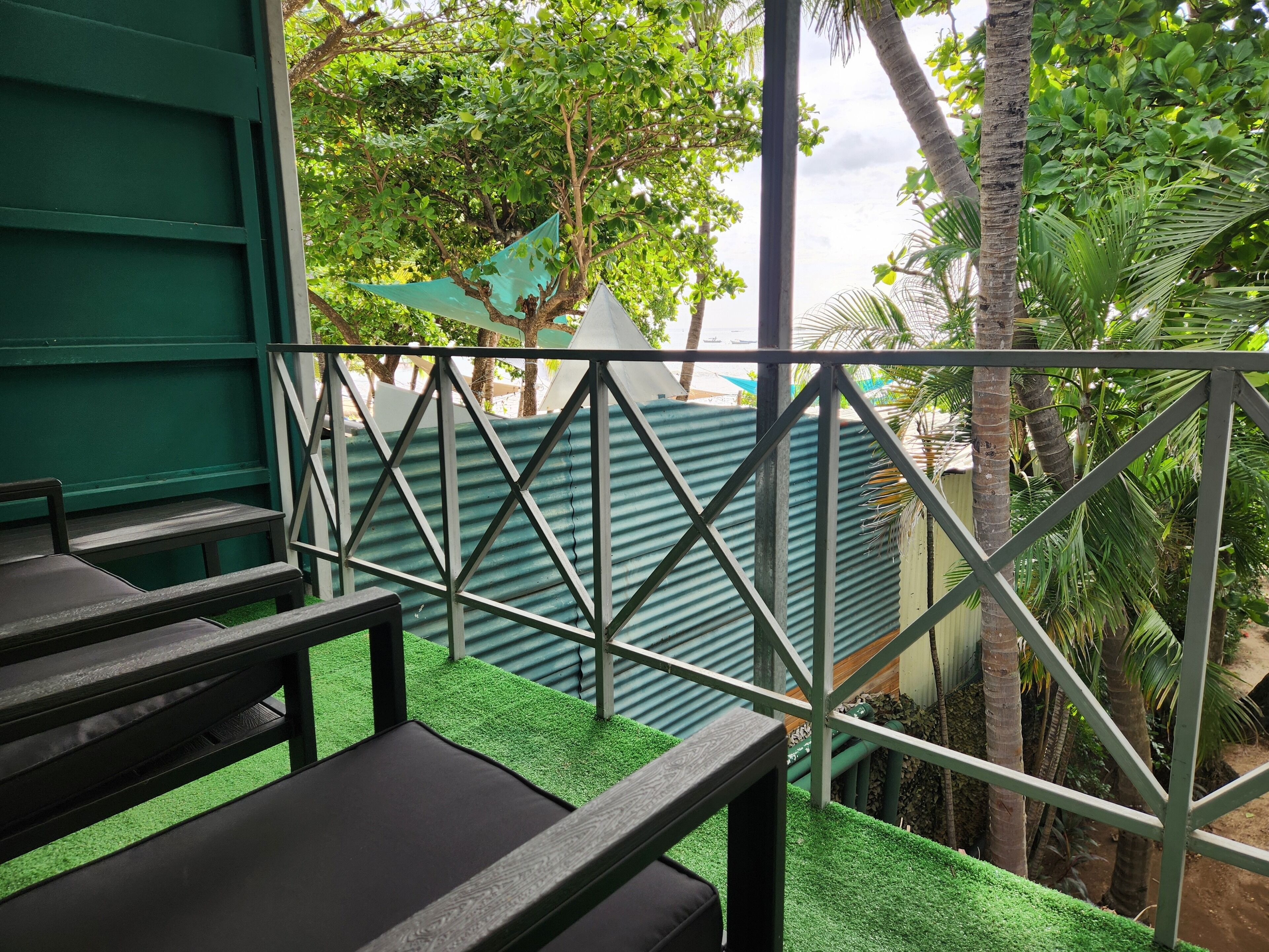 A balcony with a metal railing, green artificial grass, and a view of a building with a corrugated metal roof.