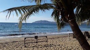 Una playa con un banco de madera, palmeras y vistas al mar y montañas lejanas.