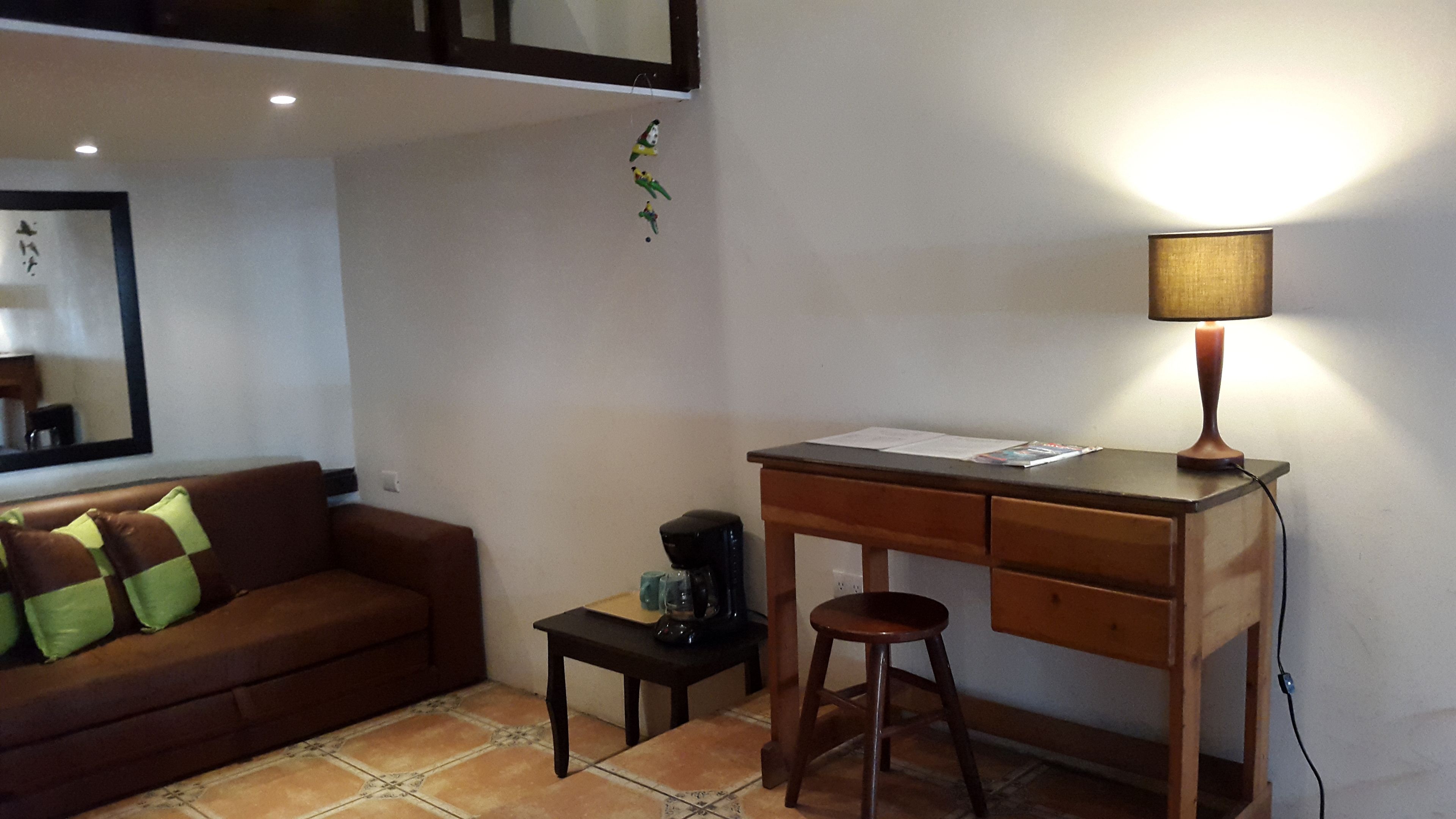 A small, furnished room with a wooden desk, a chair, a small table, a sofa with cushions, a coffee machine, and a lamp.
