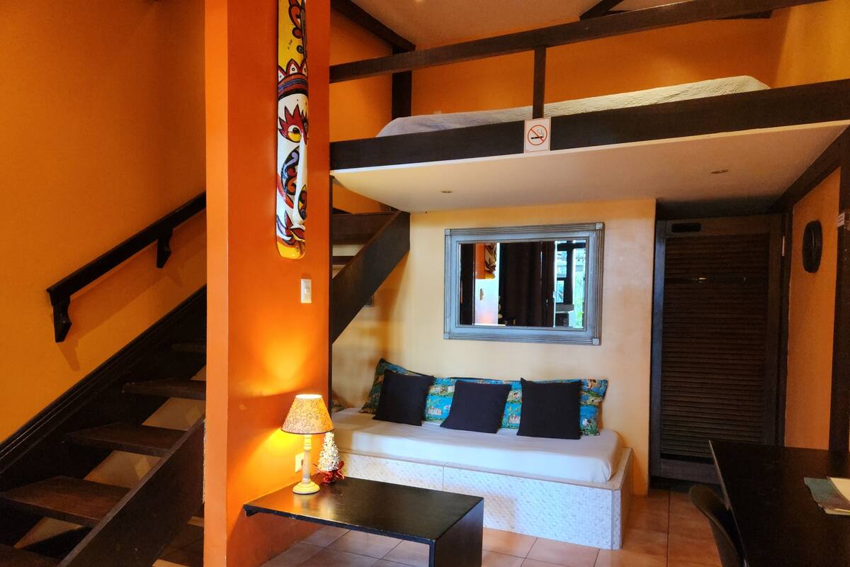 A compact living space with a staircase, a sofa with pillows, a small table with a lamp, and a window.