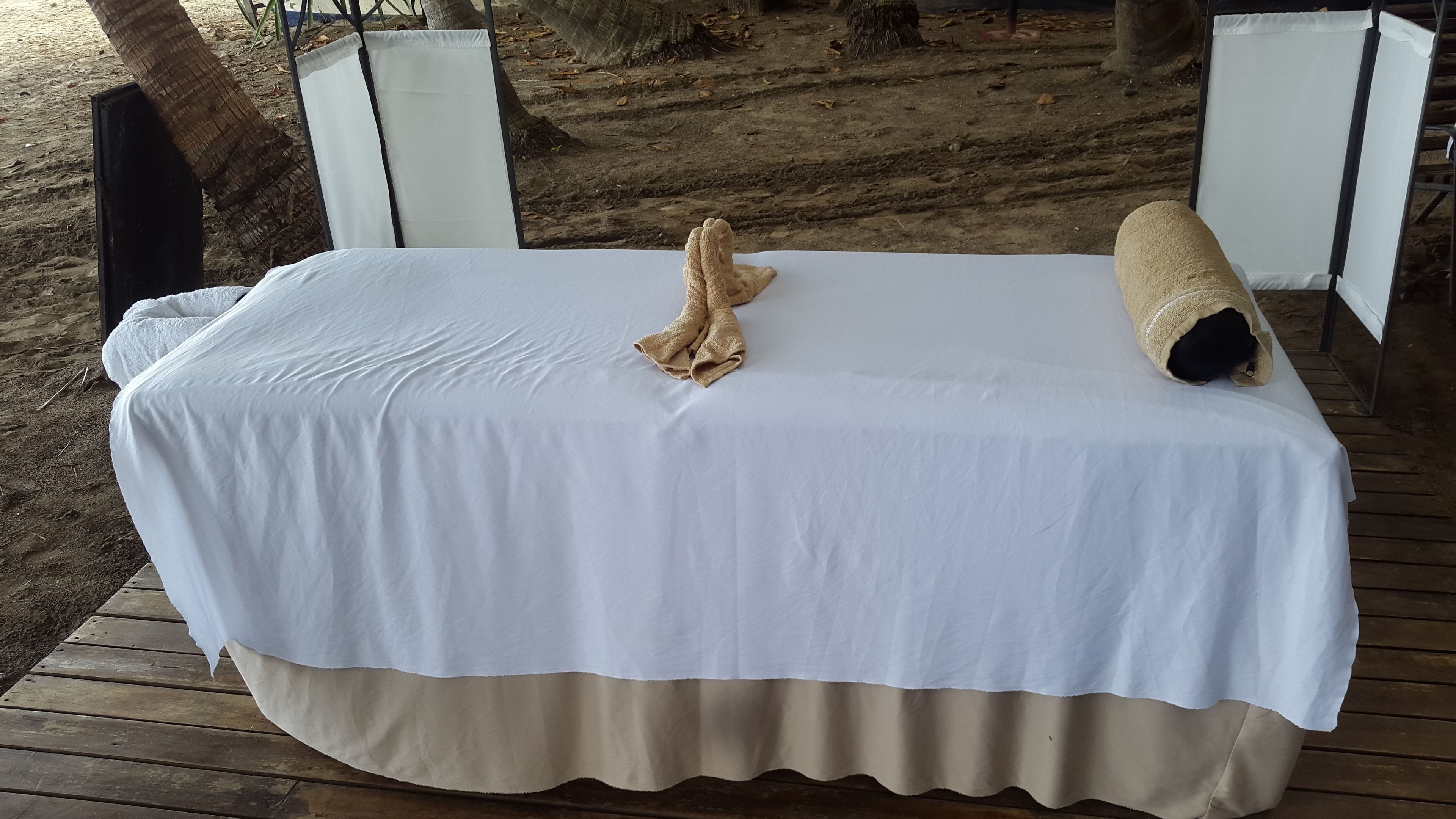 A bed with a white bedspread and beige skirt, positioned on a wooden deck with a view of a sandy area and trees.