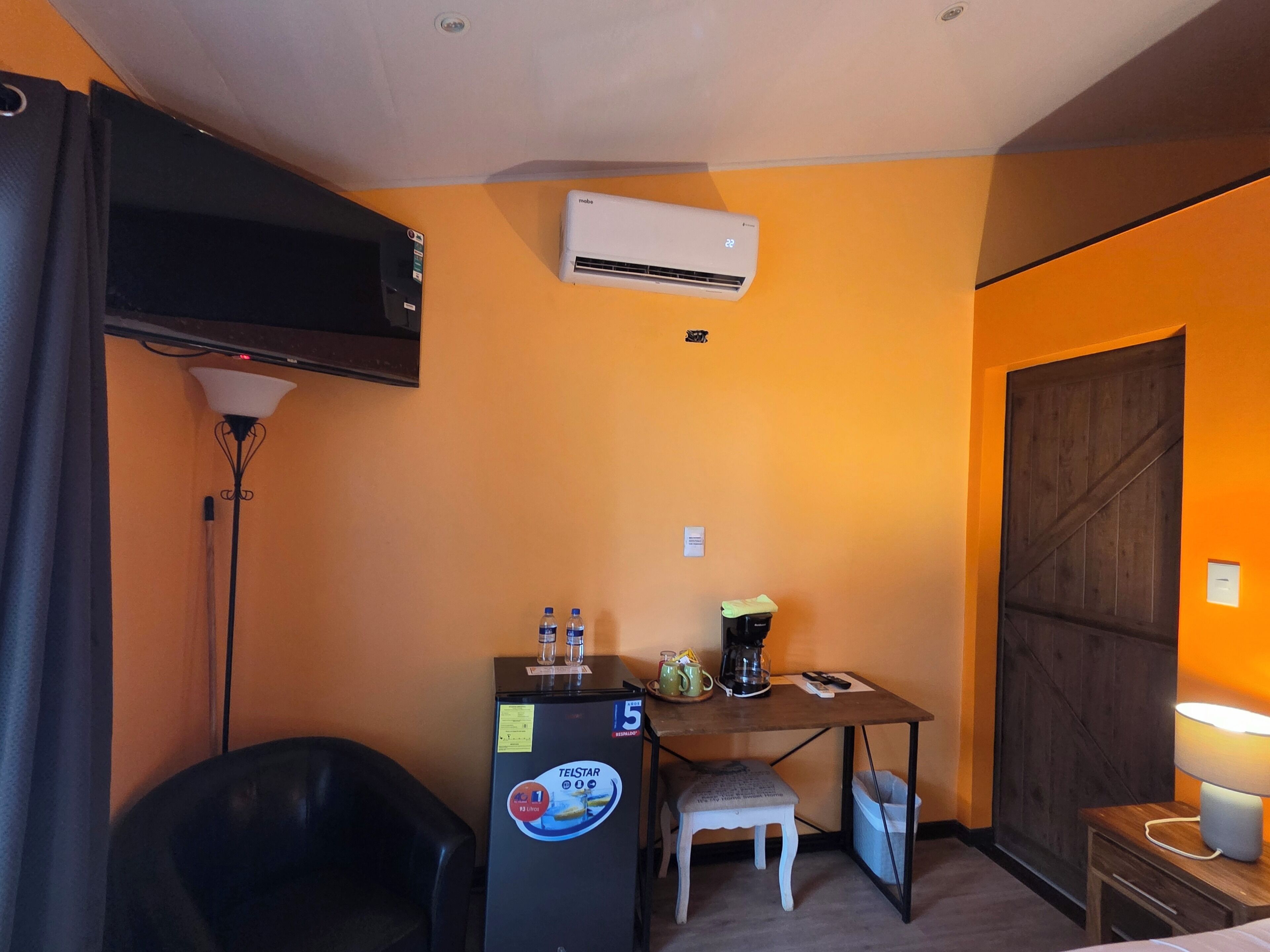 A hotel room with a bed, a desk with a chair, a refrigerator, a coffee maker, a television, and an air conditioner.