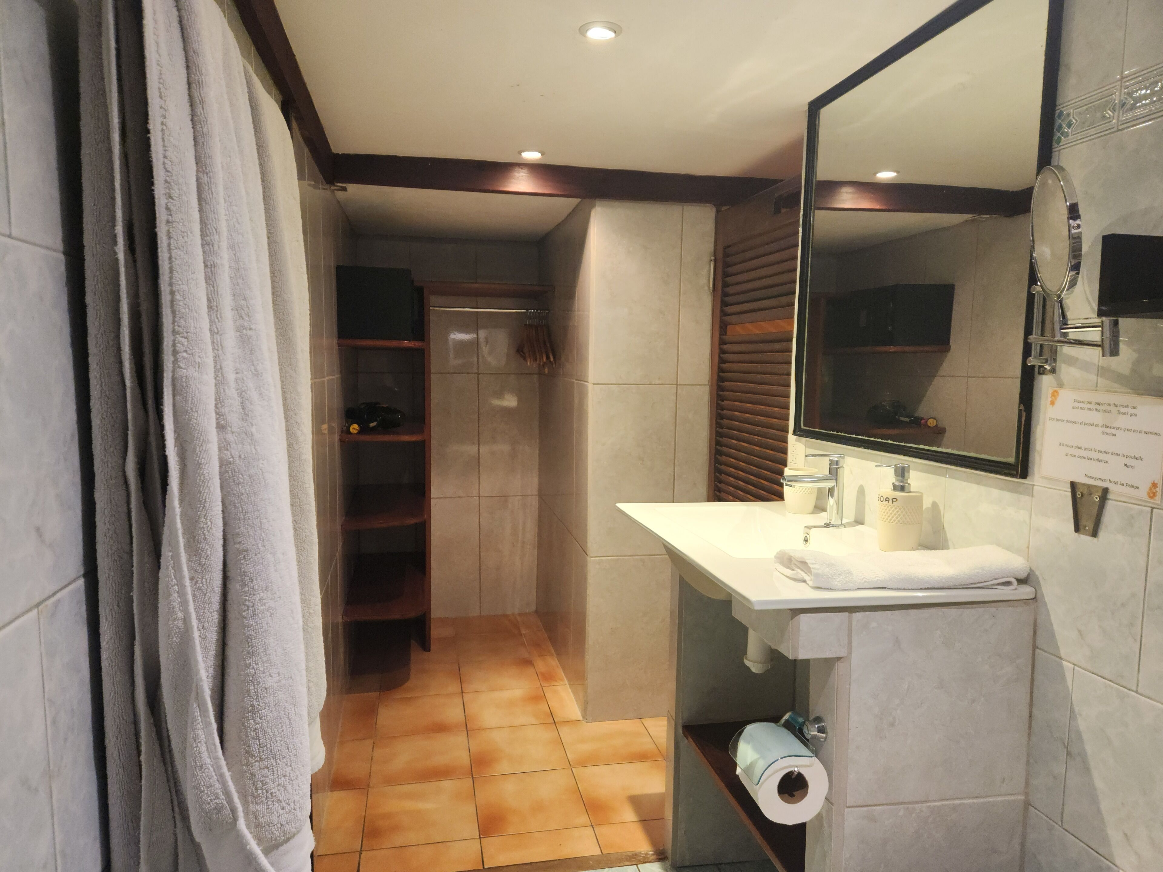 Un baño moderno con un lavamanos blanco, espejo y toallero. Se observa una zona de ducha con puerta de vidrio y un inodoro al fondo.