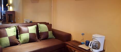 A brown leather sofa with green and brown cushions, a wooden side table with a coffee maker, and a white shelf above the sofa.