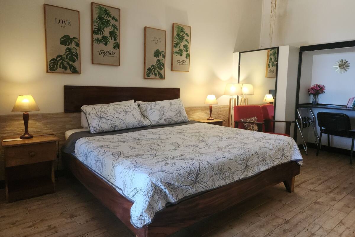 A neatly arranged hotel room with a large bed, two bedside tables with lamps, a desk with a chair, and three framed botanical prints on the wall.