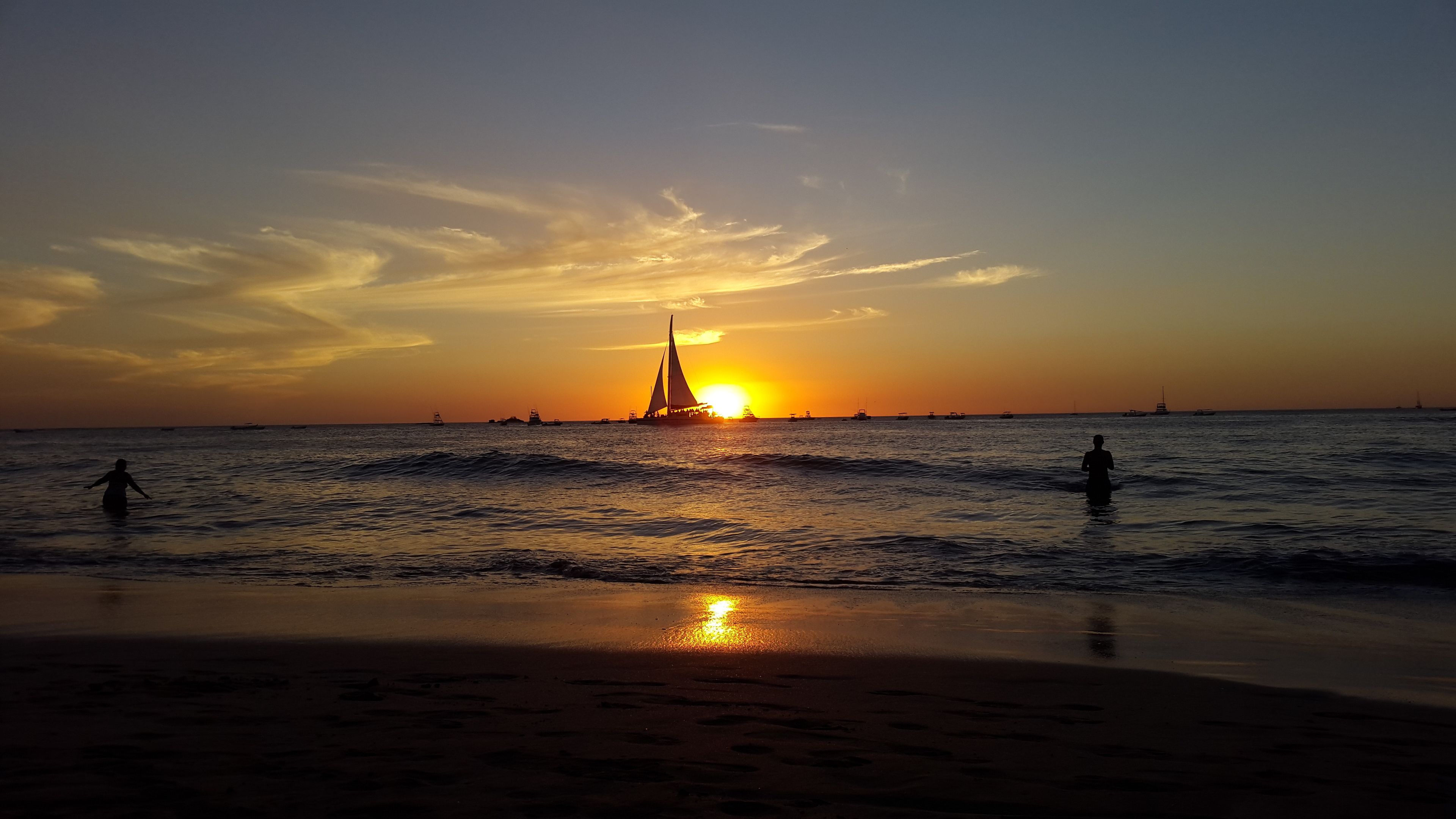 A sunset over a beach with calm waters and a sailboat in the distance.