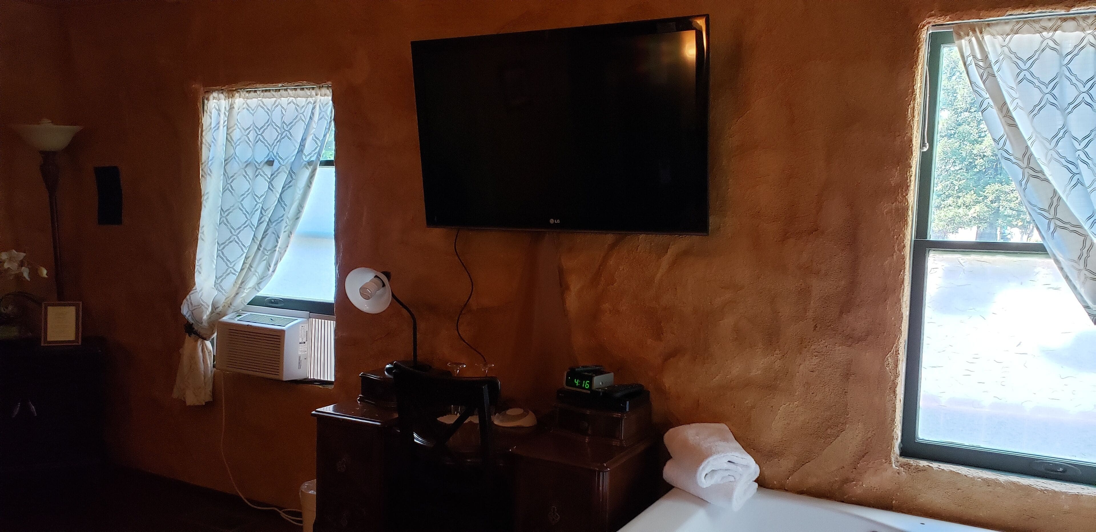 Quarto com televisão de tela plana na parede, uma escrivaninha com abajur, relógio e janela com cortinas estampadas.